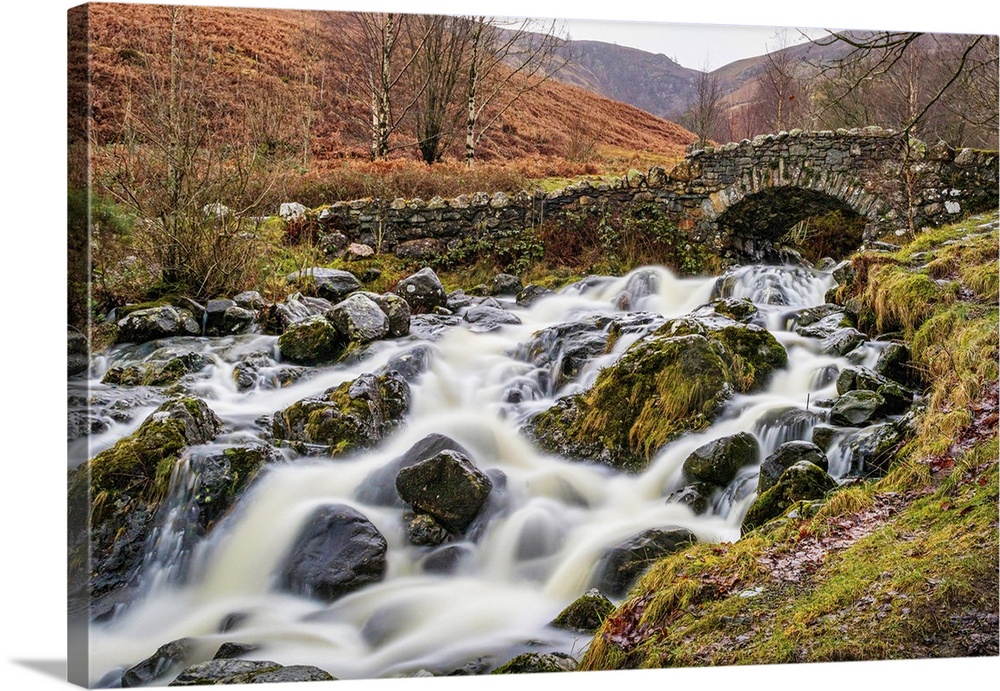 Rustic stone bridge over a flowing stream with mossy rocks in a lush, green landscape in the Lake District, Cumbria, Engla...