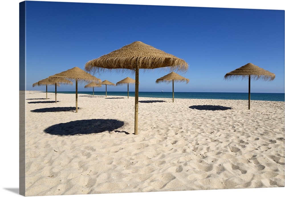 Straw umbrellas on empty white sand beach with clear sea behind, Ilha do Farol, Culatra Barrier Island, Olhao, Algarve, Po...