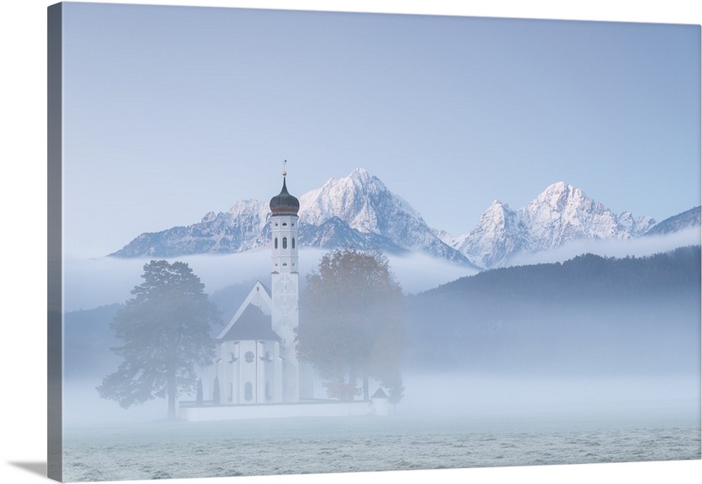 The autumn fog at sunrise frames St. Coloman Church surrounded by snowy peaks, Schwangau, Bavaria, Germany