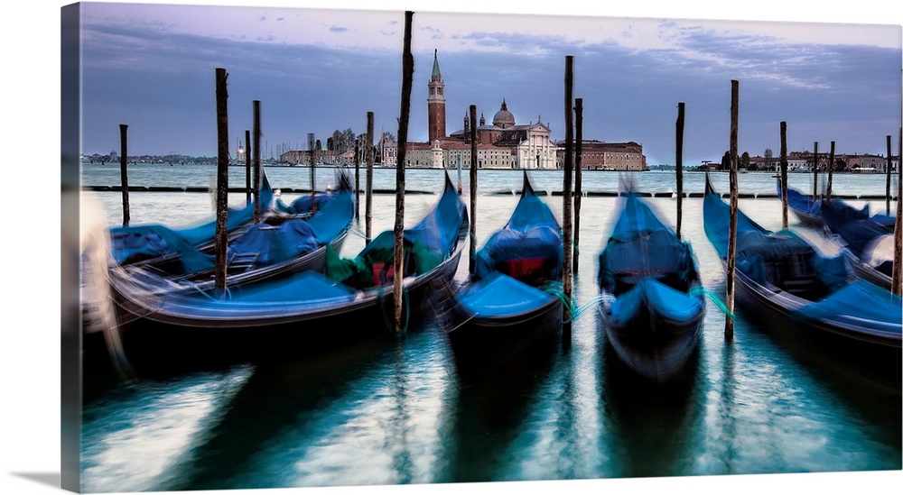 Big photograph of empty gondolas tied to poles in Venice, Italy with buildings in the background.