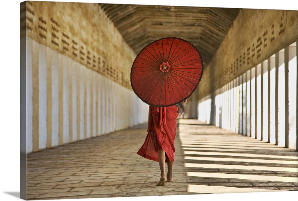 Horizontal photograph of a Monk covered from the shoulders up by an umbrella, walking down the stone hallway of a monaster...