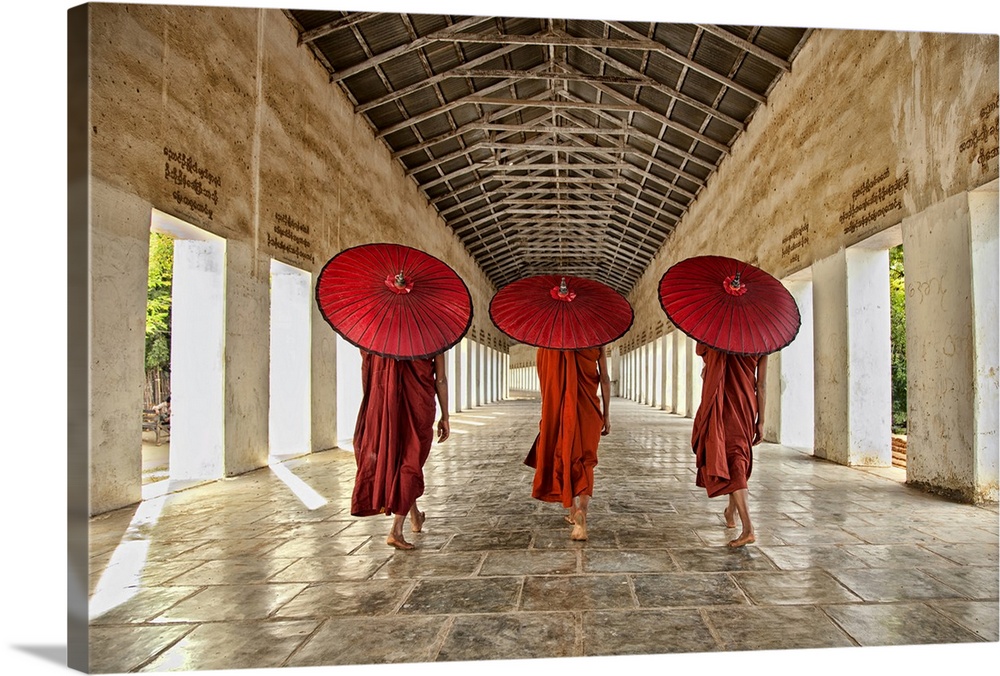 Three monks in bright red walking in a row with umbrellas down a neutral open hallway.