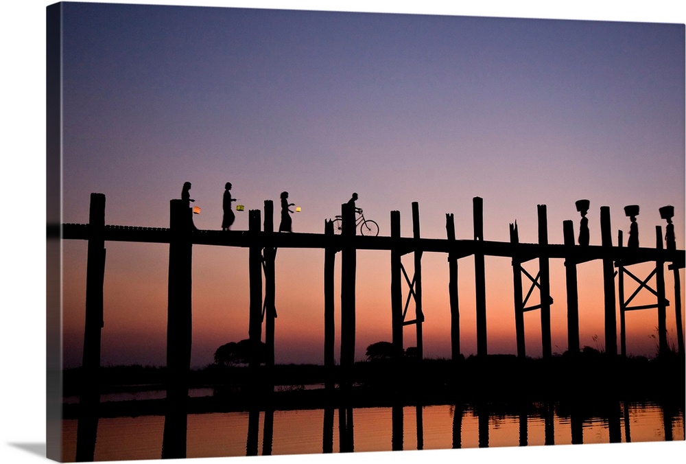 Photograph of people walking and biking across a bridge made of tall wooden beams over the ocean at dusk.  The bridge is r...