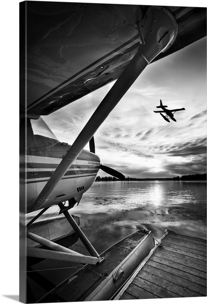 A black and white photograph taken from under the wing of a plane docked in the water while another plane has just taken o...