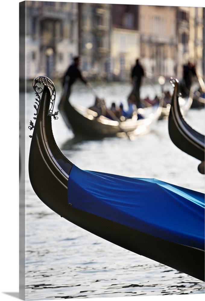 Vertical photograph of the front tip of a Venetian gondola hanging over the water.  In the background is a softly blurred ...