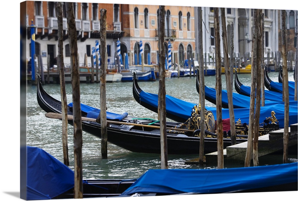 Photograph taken of a river in Venice that is lined with gondola boats that are tied up and have been covered with blue cl...