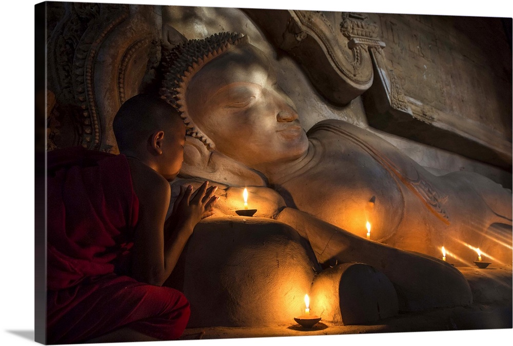 Young Burmese monk praying by candlelight by large reclining Buddha.