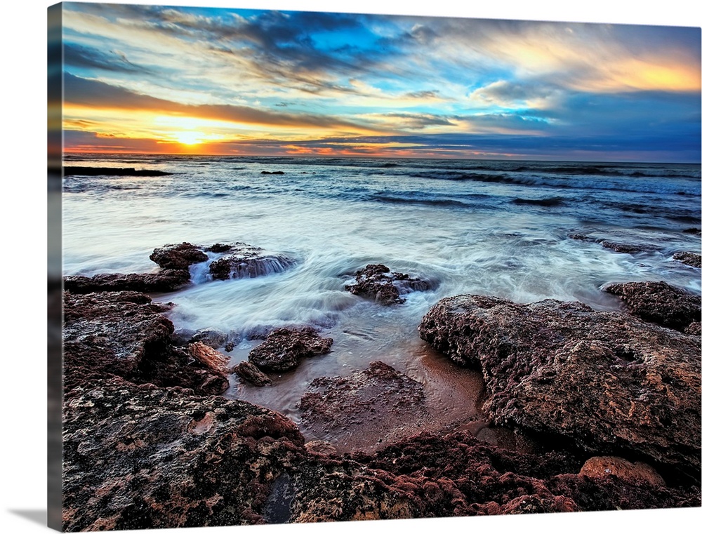 Photograph taken of the ocean that rushes up onto rocks that are pictured in the foreground. The sun is shown rising in th...