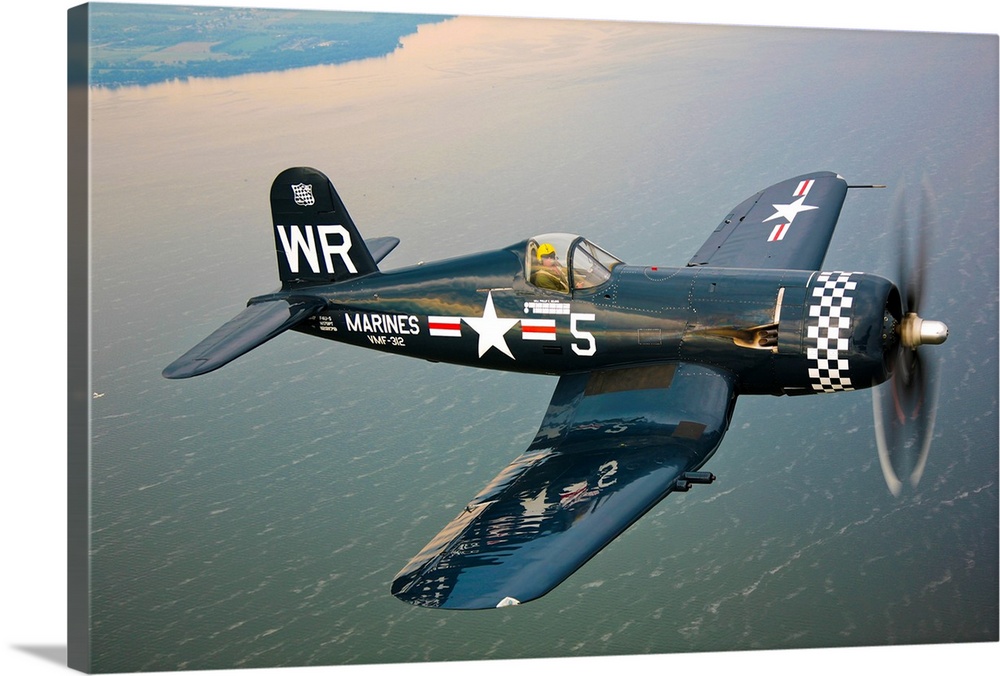 Horizontal photograph of a Vought F4U-5 Corsair in flight over a large body of water, the coastline in the distance, near ...