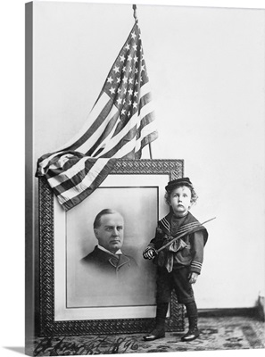 Boy in sailor uniform holding sword next to flag-draped portrait of William McKinley.