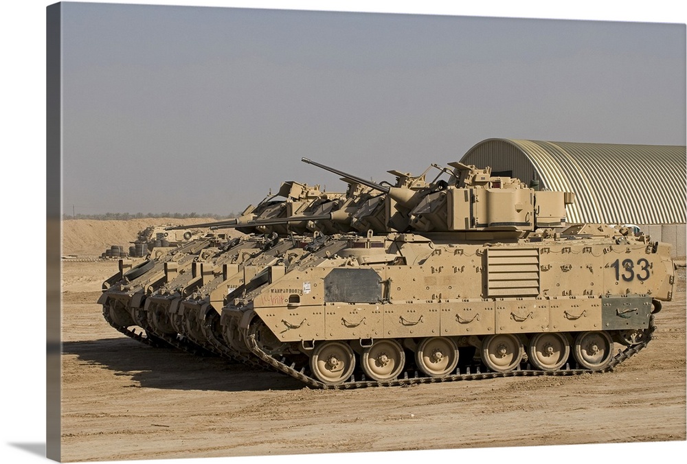 Large landscape photograph of a line of M2/M3 Bradley Fighting Vehicles cruising over a dirt landscape, a single building ...