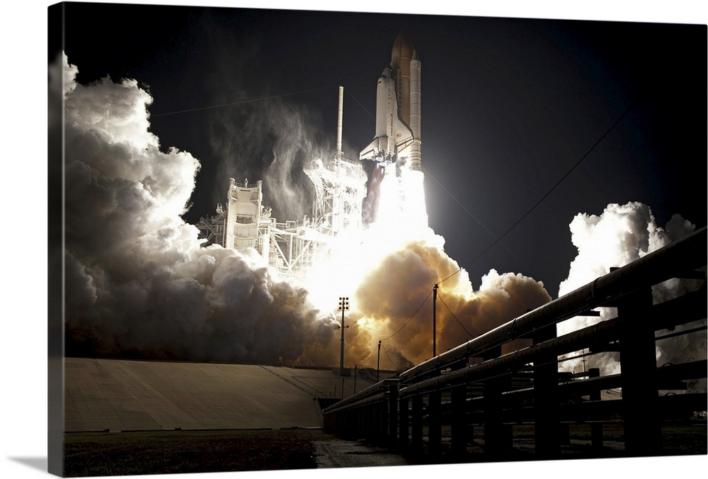 Large horizontal photograph of the space shuttle Endeavour taking off from the launch pad at night, leaving behind a brigh...