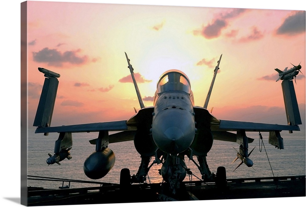 Military photograph of a jet aircraft on the deck of a carrier ship, with wings folded up and the sun rising behind the co...