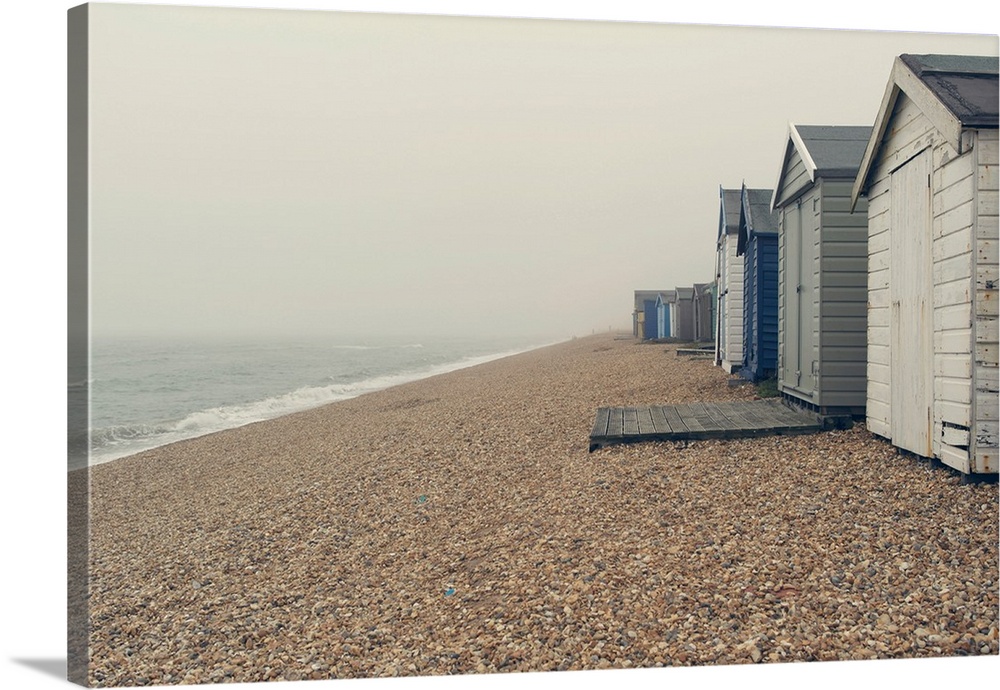 A photograph of a pebbled beach with a row of beach huts on an overcast day.