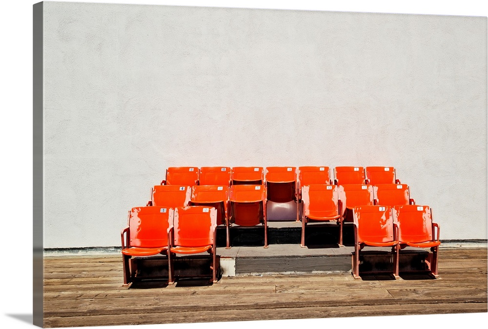 Photograph of three rows of bright orange theater seats against a plain wall and wooden floor.