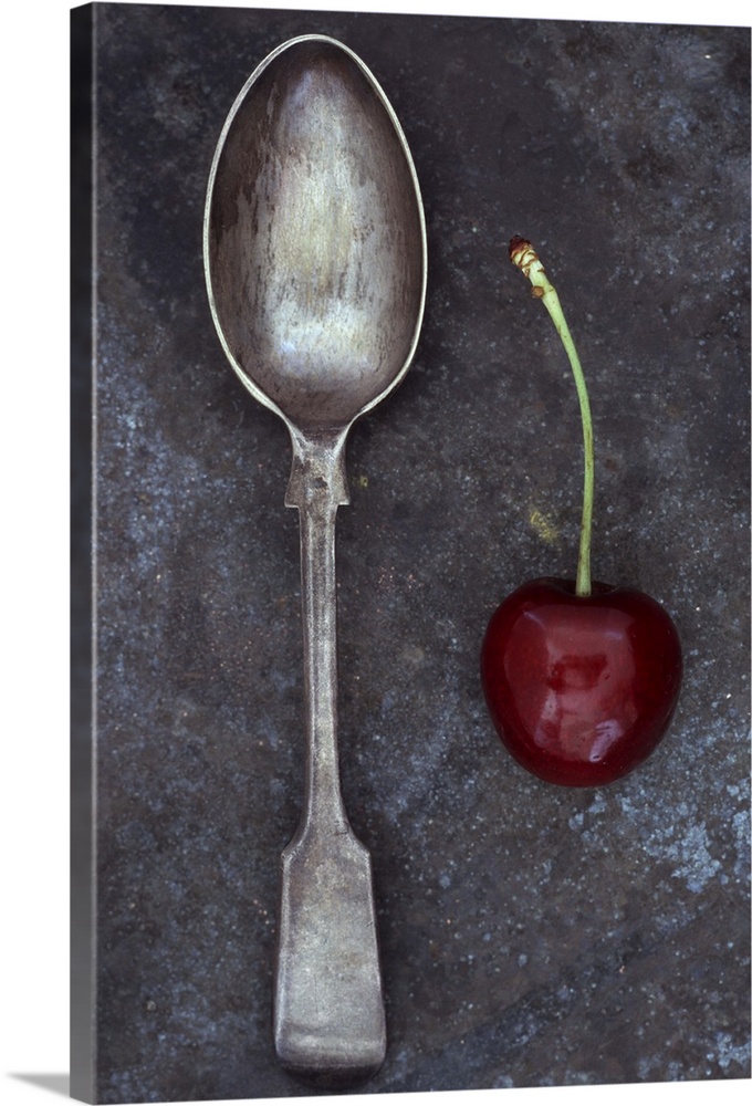 Antique tarnished silver teaspoon lying next to single dark red cherry with stalk on tarnished metal
