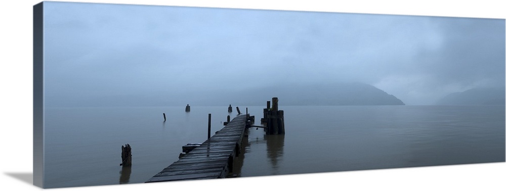A storm brews over a still lake as a rundown pier still stands in this foggy serene photograph.