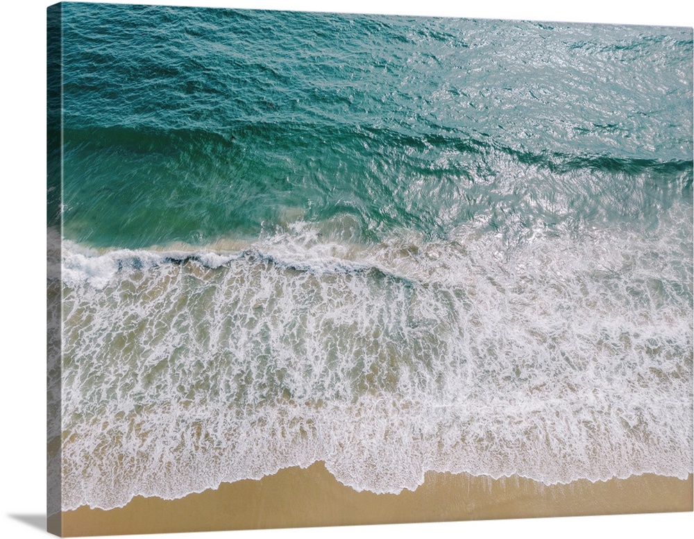 An overhead photograph of clear blue waters fading into white foam lapping a smooth sandy beach.