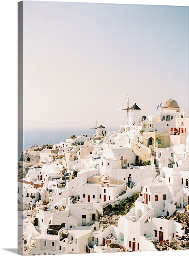 A photograph of the iconic white stucco buildings and windmills that are characteristic of the hillside town of Santorini,...