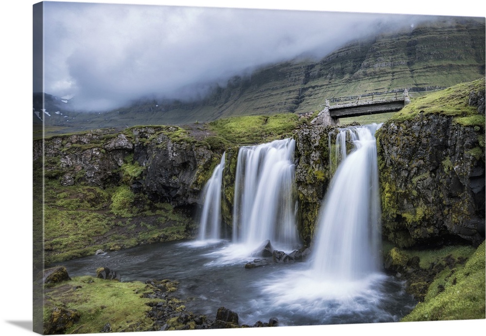 Photograph of waterfalls from a river pouring into a small pool with a worn bridge and mountains obscured by fog in the ba...