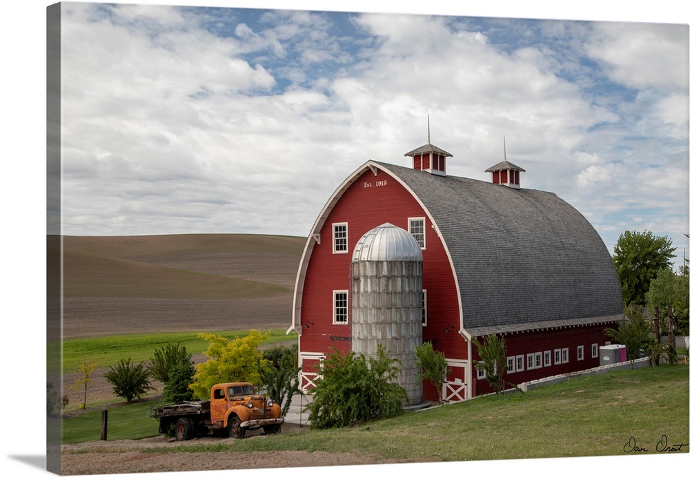 A photograph of a large red barn and grain silo with a vintage orange truck parked out front
