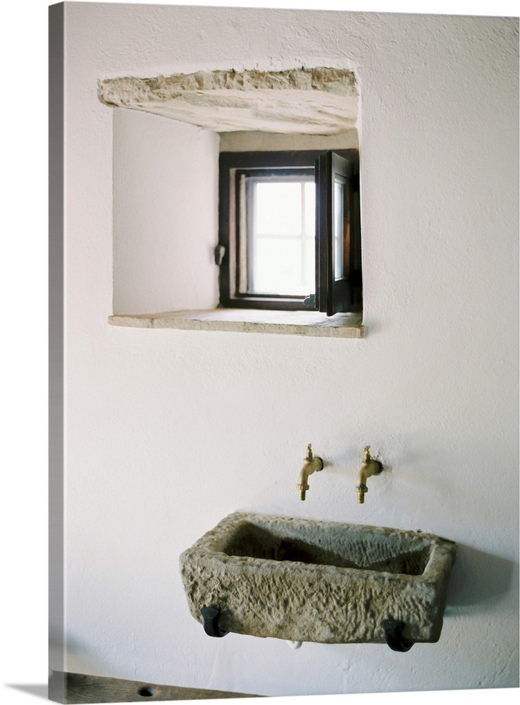 A photograph of a rustic stone wall basin underneath a small window in a white stucco mediterranean dwelling.