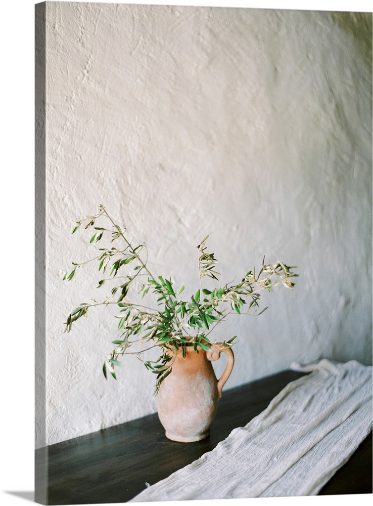 A photograph of olive branches in a clay jug in a simple indoor mediterranean setting.